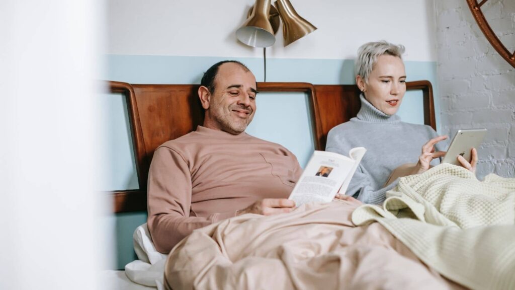 Homem lê livro deitado em sua cama enquanto esposa deita ao seu lado lendo na biblioteca digital da POLI USP PRO em um tablet.