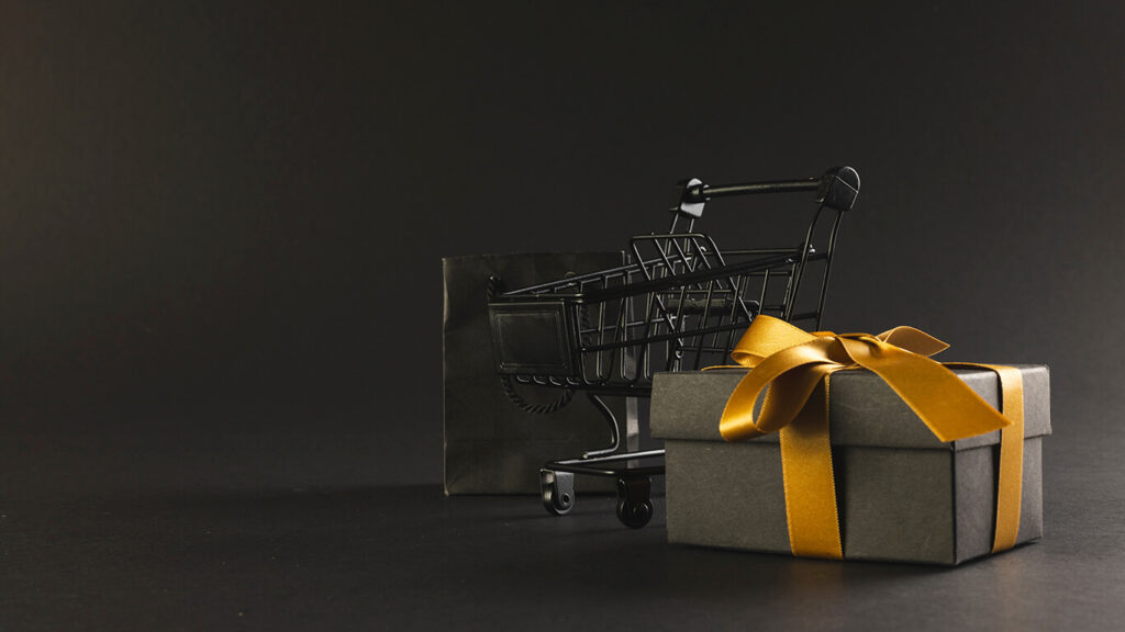 Um carrinho de mercado em miniatura, ao lado de um presente e um laço, em um cenário cinza.