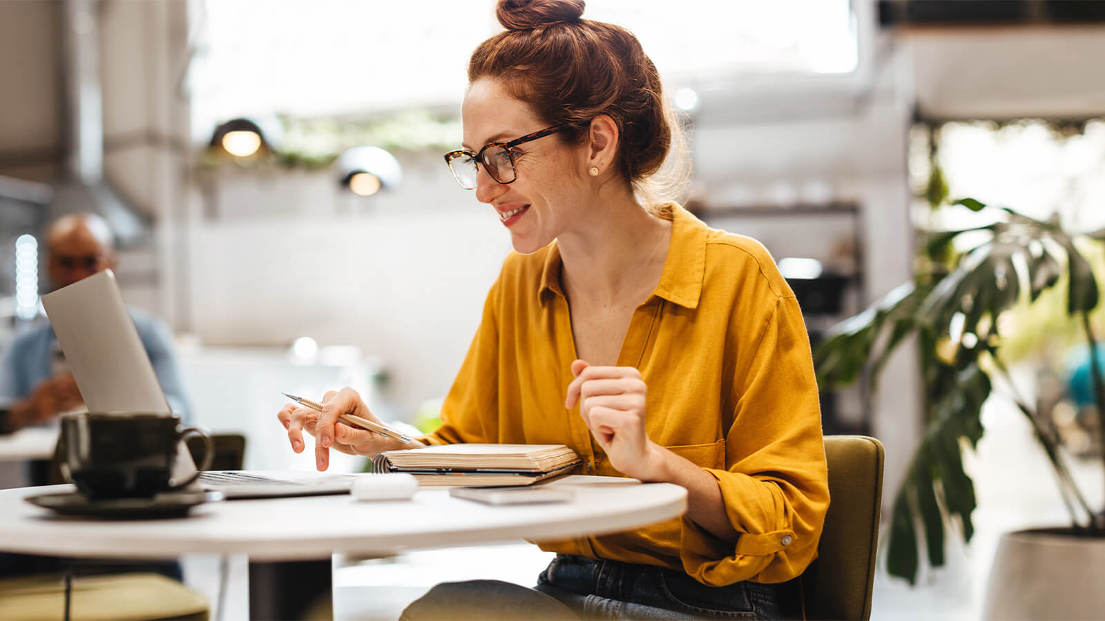 Uma mulher com camisa amarela se senta de frente para uma mesa, onde está apoiado um notebook e ela parece estudar, em relação à importância da pós-graduação no mercado de trabalho.