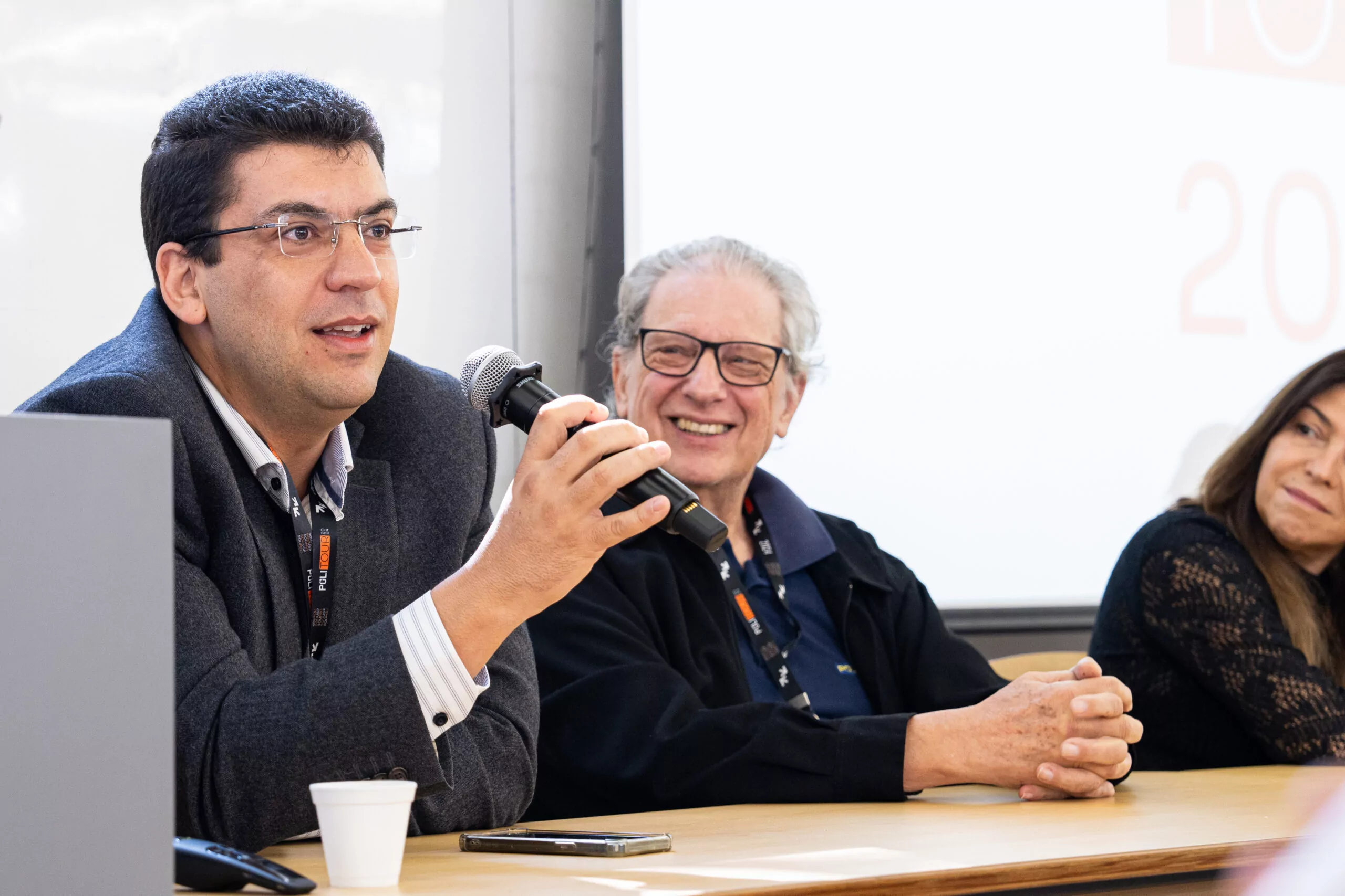 foto mostra professor Daniel com microfone em primeiro plano, a esquerda. No meio vemos outros professor sorrindo e a direita professora Marly.